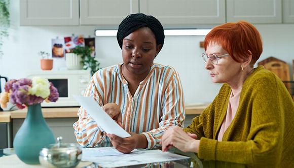 younger lady sitting with older lady at kitchen table looking over documents